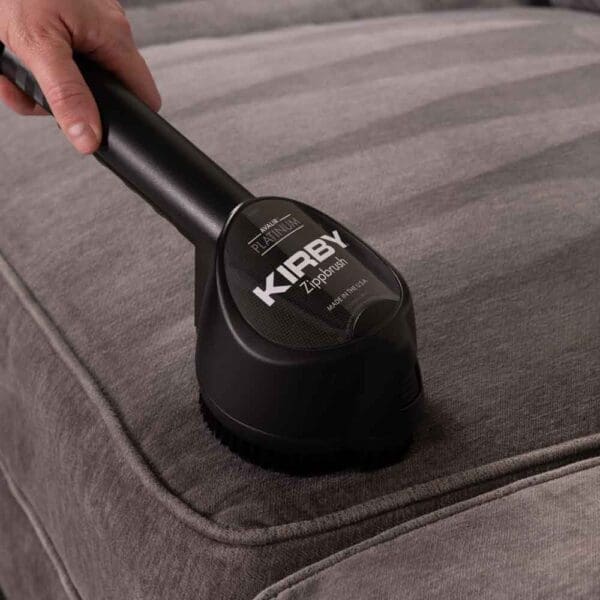 Hand using black Kirby Zippbrush on a gray couch.
