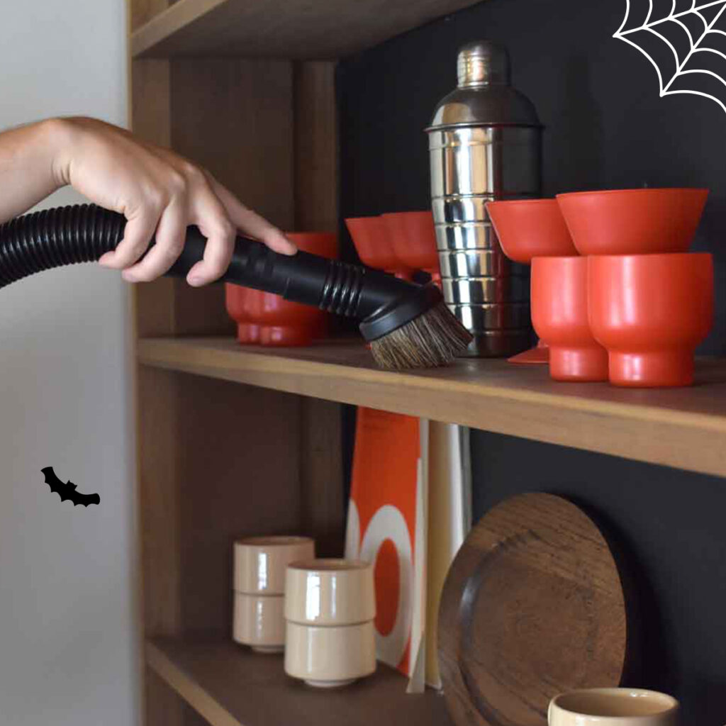 Left hand holding vacuum hose with brush attachment cleaning shelves with Halloween-themed dishware.