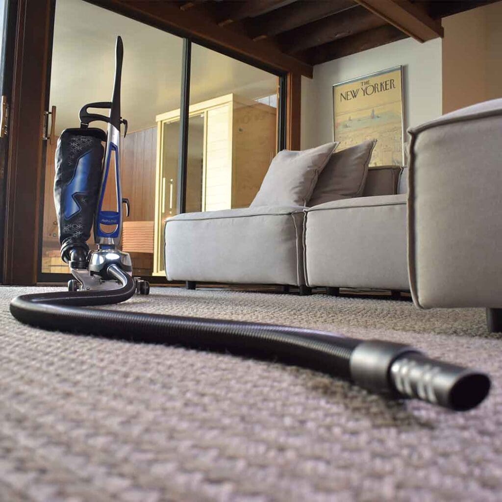 A Kirby vacuum cleaner with it's hose attached on the floor in a living room with a couch and a chair in the background